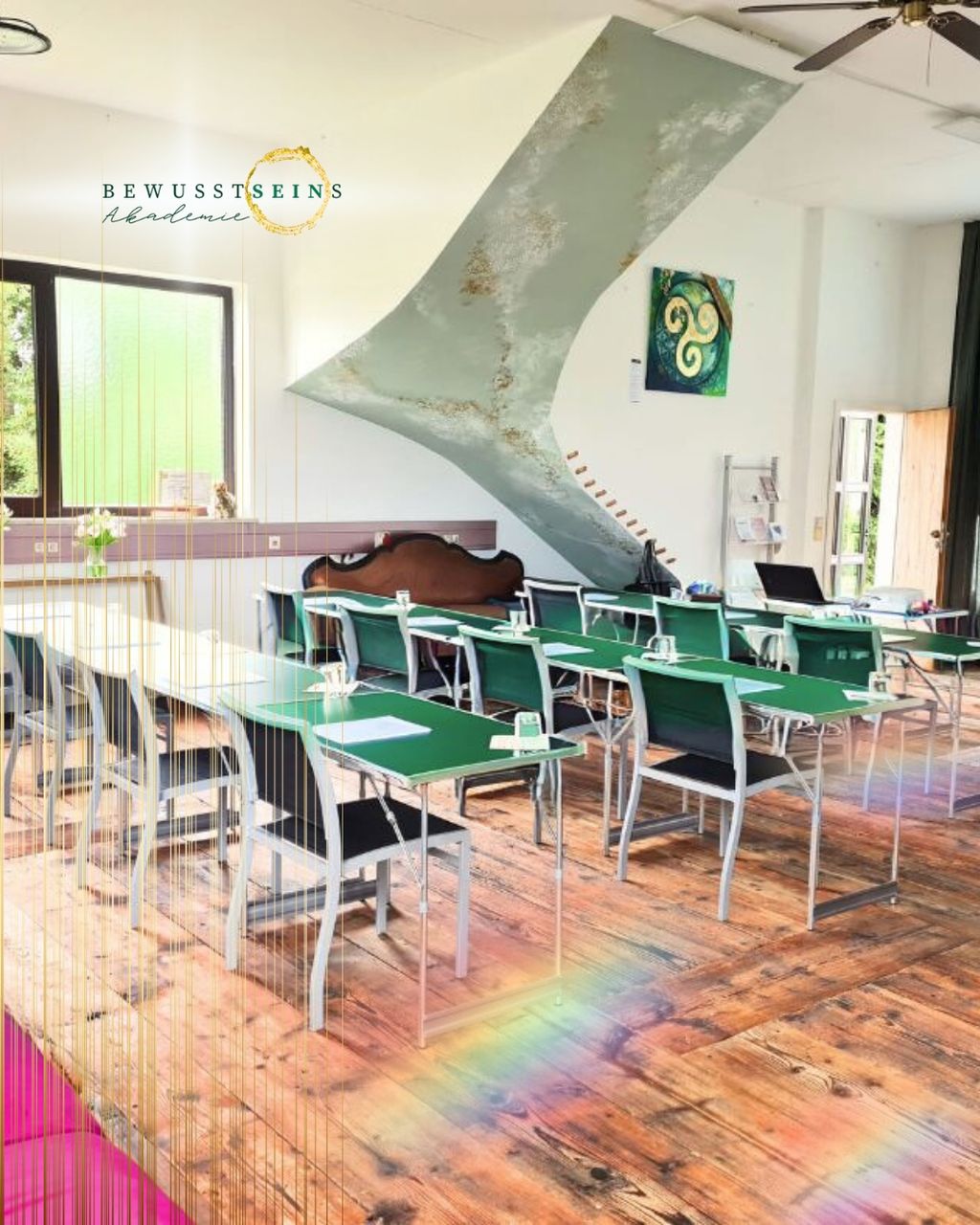 Klassenzimmer der Bewusstseins Akademie mit grünen Tischen und Stühlen, Holzboden, großen Fenstern und moderner Deckenbeleuchtung in einem hellen Raum.